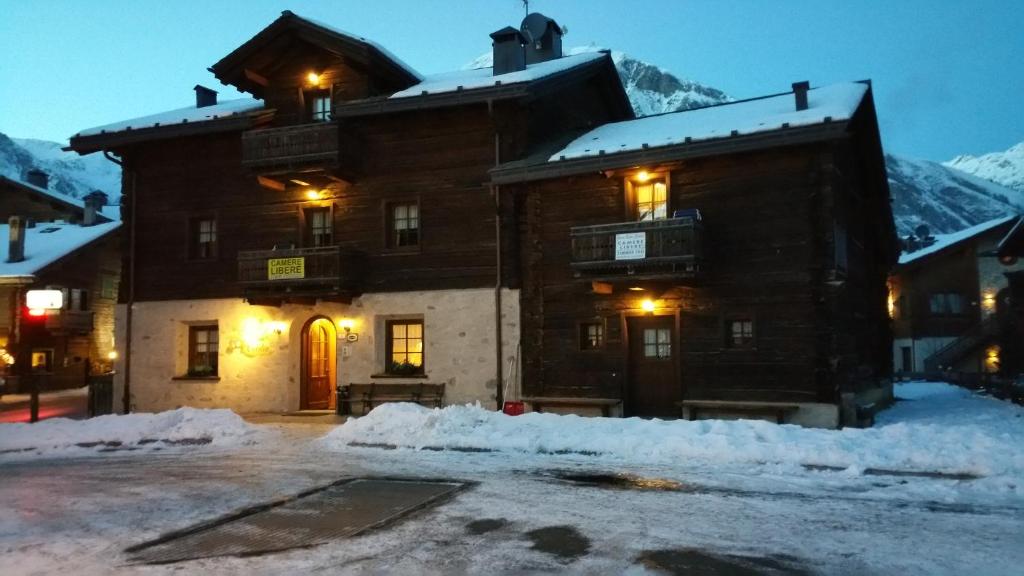 Tradicionāla kalnu māja ar koka fasādi, apņemta ar sniegu un kalnu ainavām. Siltas gaismas atspīdums no logiem rada mājīgu atmosfēru.