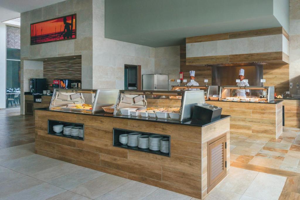 Mūsdienīgs restorāna interjers ar bufetes galdu, redzami šefpavāri un ēdienu izvietojums. Gaišas krāsas un koka elementi rada viesmīlīgu atmosfēru.