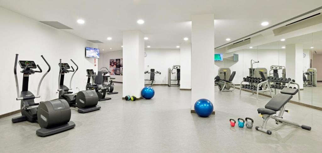 Mūsdienīgs sporta zāles interjers ar dažādiem trenažieriem, hantelēm un vingrošanas riepām. Telpa ir gaiša un plaša, nodrošinot komfortablu vidi treniņiem.