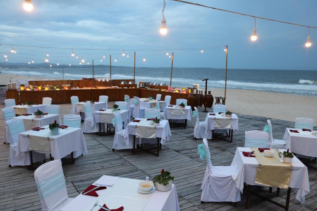 Pludmales restorāna terase ar galds un krēsli, dekorēti ar ziediem, un apgaismojums, kas radīts vakara atmosfērai. Jūras skats un viļņi veido relaksējošu fonu.