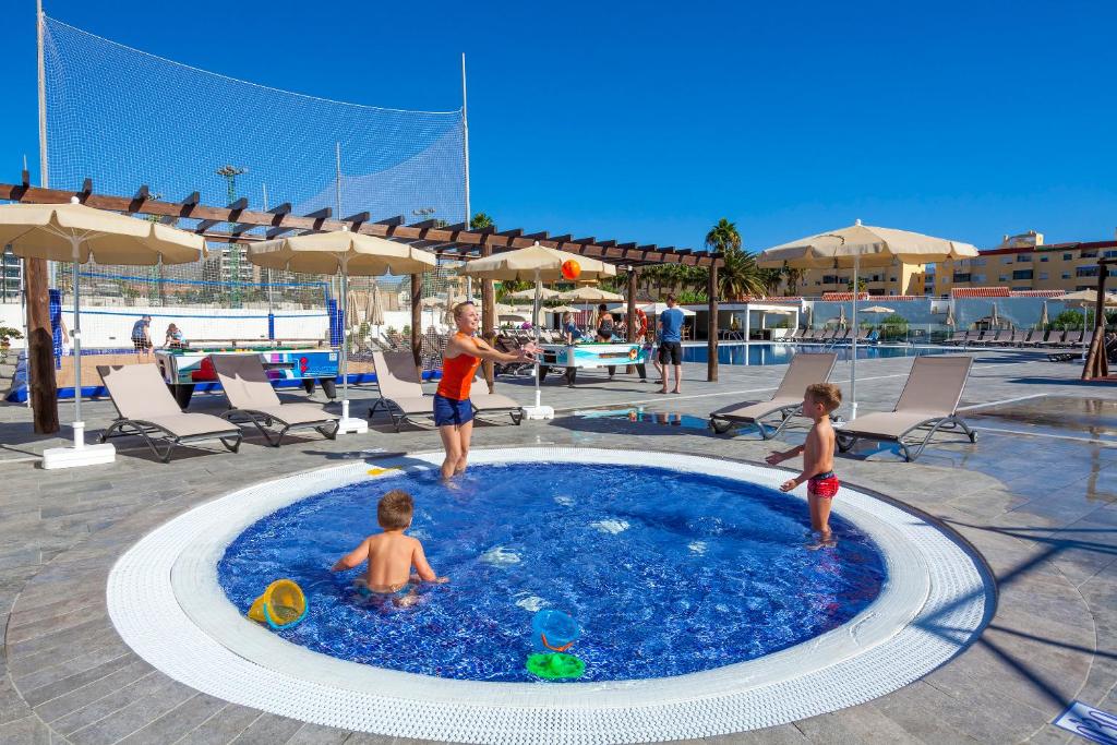 Bērni un pieaugušie izbauda ūdens prieku baseinā, kamēr saule spīd skaidrā debesīs. Ap baseinu atrodas sauļošanās krēsli un saulessargi, radot relaksējošu atmosfēru.