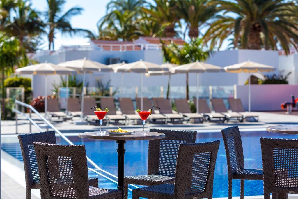 Peldbaseins, kurā redzami sarkanie kokteiļi, sēžot uz galdiem, un saulessargi, kas aizsargā no saules, veido relaksējošu atmosfēru. Apkārt ir ērtas sauļošanās vietas un zaļie palmas, kas piešķir tropisku noskaņu.