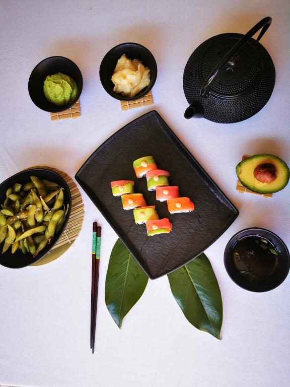 Svaigas suši šķēles uz melnas plāksnes, kopā ar edamame, wasabi, ingveru un tējas podiņu. Galda kompozīcija ietver arī avokado un koka pārus.