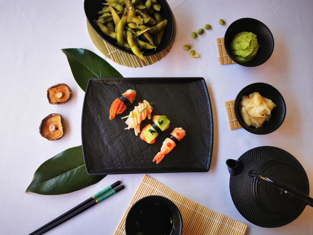 Svaiga sushi uz melnas plāksnes kopā ar edamame, wasabi un marinētiem ingveriem, papildināta ar tējas katliņu un koka paklājiņu.