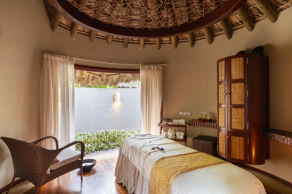 Relaksējoša spa telpa ar dabīgām koka sijām, gultu un ērtu atpūtas krēslu. Logi ar aizkariem nodrošina dabīgu apgaismojumu un skatu uz dārzu.