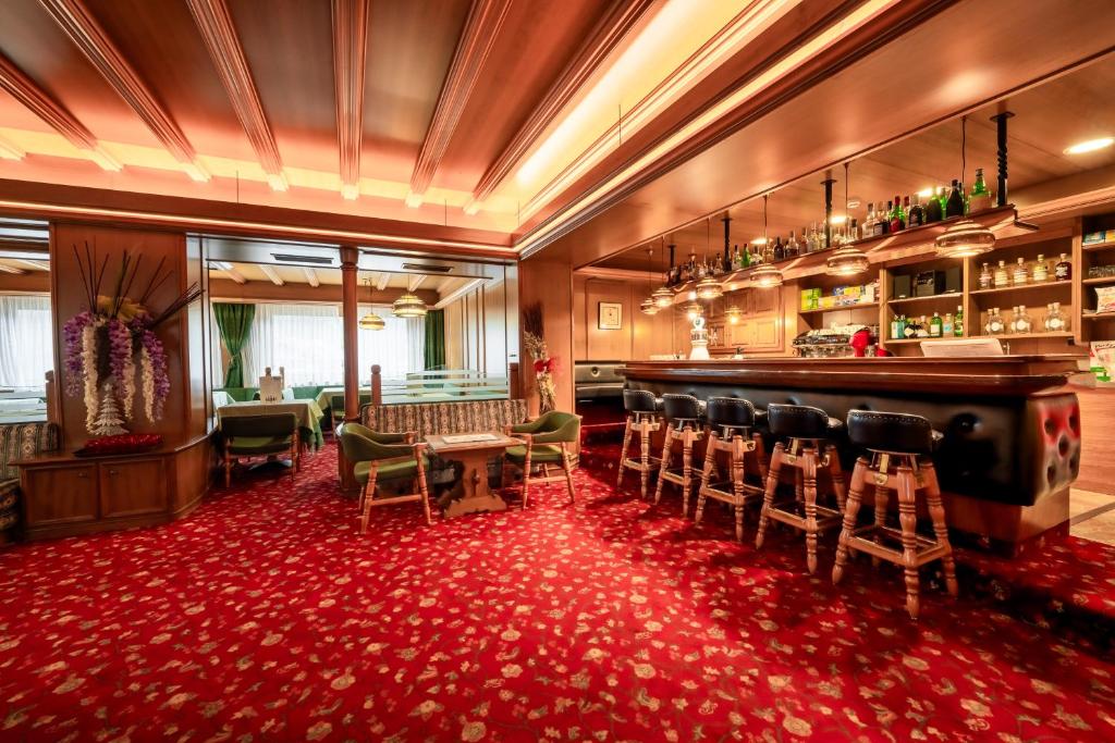 Eleganta bāra iekšskats ar koka apdari un sarkanu paklāju, kurā redzami bāra krēsli un plaukti ar dzērieniem. Gaisma ir silta, radot mājīgu atmosfēru.