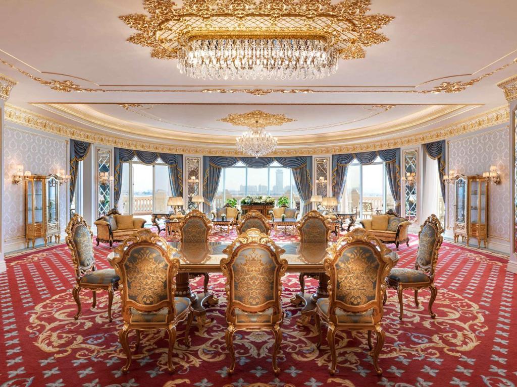 Luksusīgs viesistabas interjers ar greznu galdu un krēslām, zeltītiem elementiem un plašām logu atverēm, caur kurām redzams pilsētas skats. Eleganta sarkanā paklāja un izsmalcinātas apgaismojuma ķermeņi piešķir telpai īpašu šarmu.