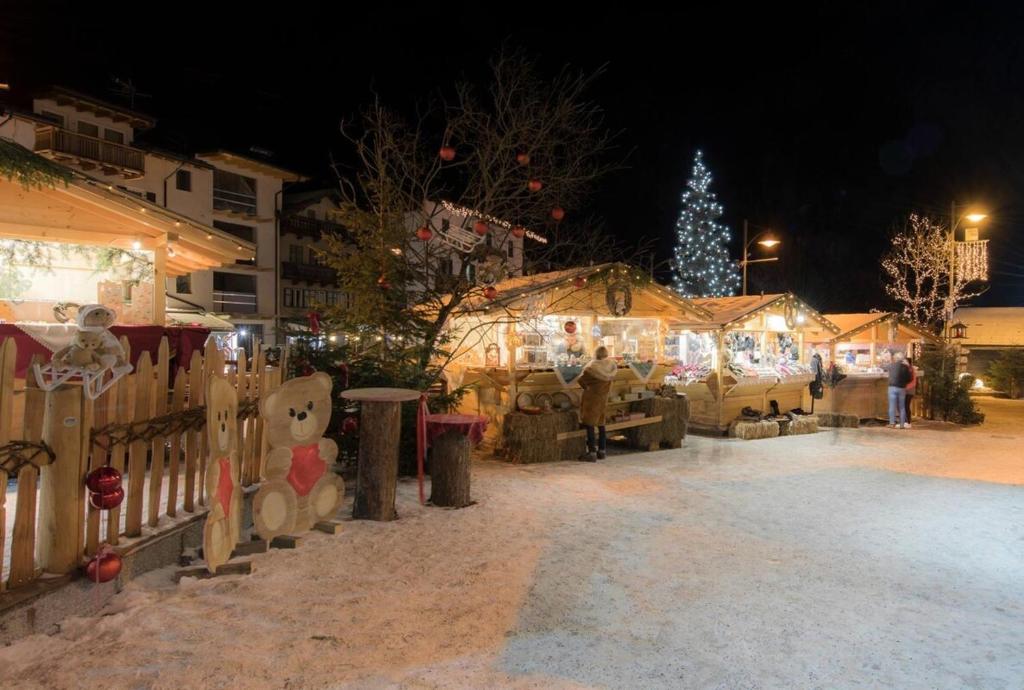 Ziemassvētku tirgus ar dekoratīvām koka būdām un gaismiņām, sniegots fons, un egles ar rotājumiem. Apkārt cilvēki izbauda svētku atmosfēru.