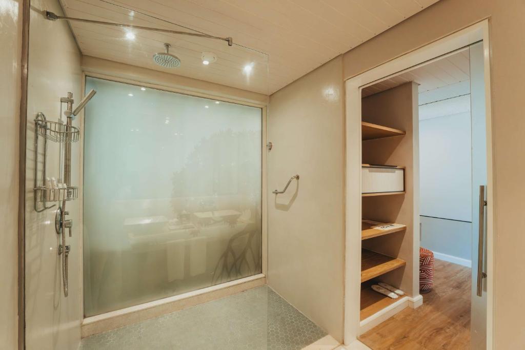 Mūsdienīga dušas telpa ar caurspīdīgu stikla sienu un stilīgu apgaismojumu, kas piešķir telpai gaisīgumu un modernu izskatu. Uzglabāšanas plaukti blakus dušai nodrošina ērtu lietu organizēšanu.
