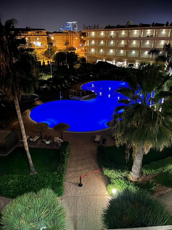 Nakts skats uz zilo baseinu viesnīcas dārzā, ko ieskauj palmas un apgaismotās ēkas. Baseins ir izveidots unikālā formā un rada relaksējošu atmosfēru.