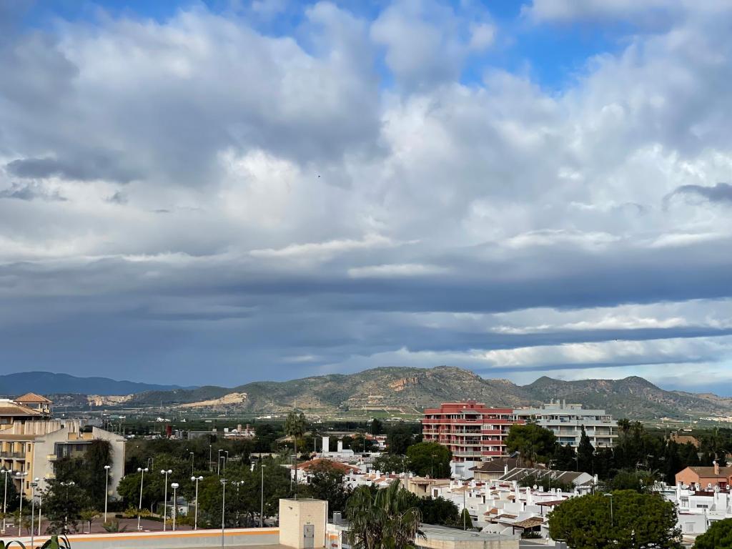 Skats uz kalniem un pilsētas ainavu ar mākoņainu debesu fonu, kur redzami dažādi dzīvojamie un komerciālie objekti.