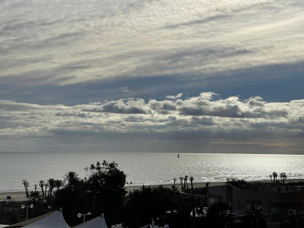 Jūras skats ar viļņiem un burājošu laivu, fons ir mākoņains debesis un saules gaisma, kas atspoguļojas ūdenī. Pludmale un palmas pie horizonta.