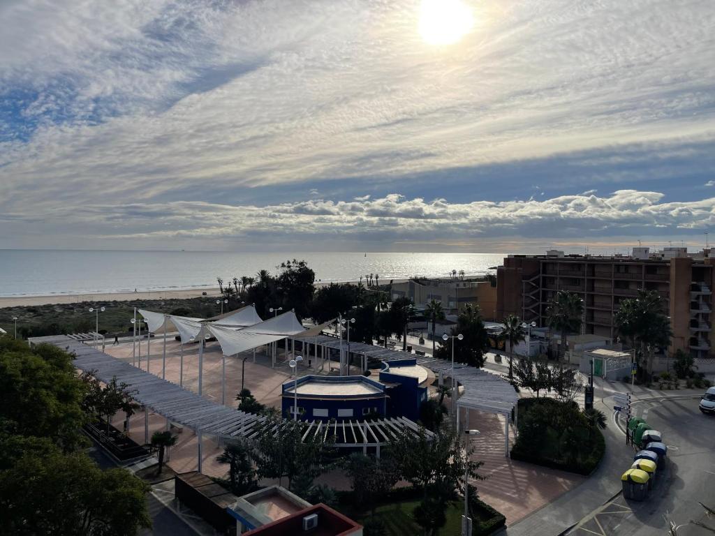 Skats uz pludmali ar sauli un mākoņiem, priekšplānā ir moderns laukums ar baltiem nojumes elementiem un zaļiem kokiem, blakus ir ēkas un automašīnas.