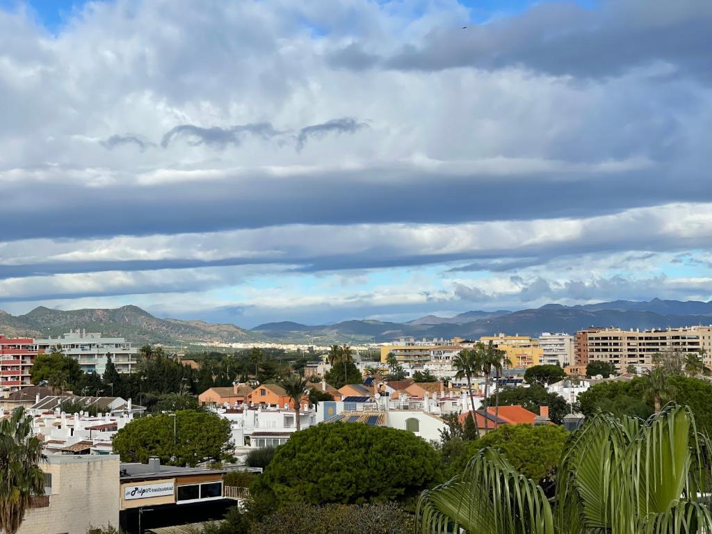 Skats uz kalniem un pilsētas ainavu ar mājām, zaļiem kokiem un mākoņainām debesīm.