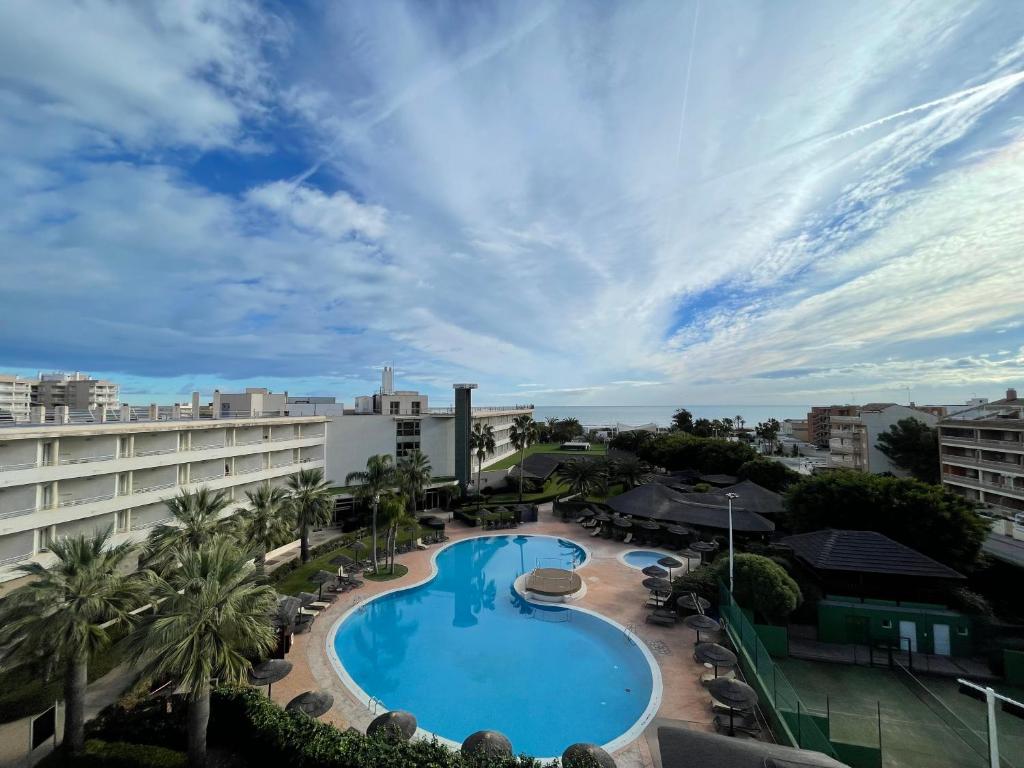 Viesnīcas ārējais skats ar baseinu un palmām, fons ir zila debess un jūras krasts. Ideāla vieta atpūtai un relaksācijai.