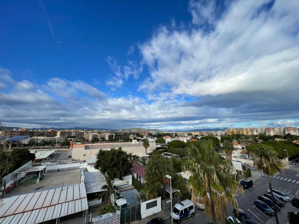 Pilsētas ainava ar mākoņainu debesu fonu, kur redzami daudzi ēkas un zaļi koki. Skats uz modernu apkaimi ar asfaltētiem ceļiem un automašīnām.