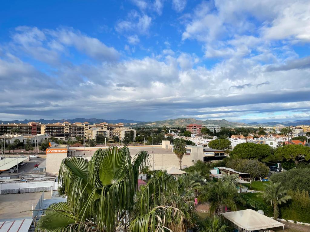 Skats uz pilsētas ainavu ar modernām ēkām un kalnu fona, klātas ar zemu zaļu krūmu un palmām. Debesis ir daļēji mākoņainas, radot mierīgu atmosfēru.