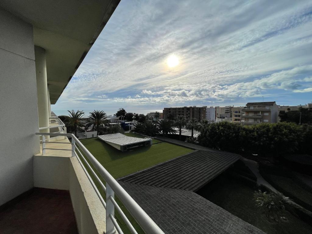 Skats no balkona uz pilsētas ainavu ar sauli, kas spīd cauri mākoņiem, un zaļu zālienu, kas ieņēmis daļu no ainavas.