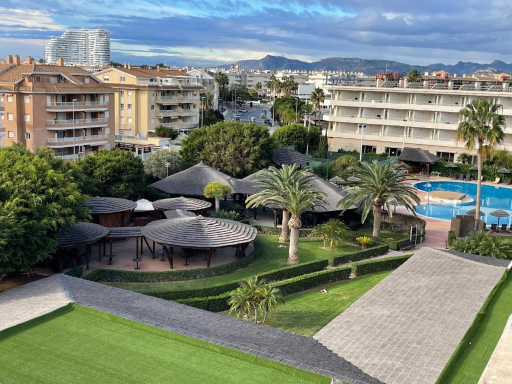 Ainava ar viesnīcu kompleksu un peldbaseinu, ieskautu zaļām palmu un augļu kokiem, nodrošina relaksāciju un atpūtu. Fonā redzami kalni un pilsētas ainava.