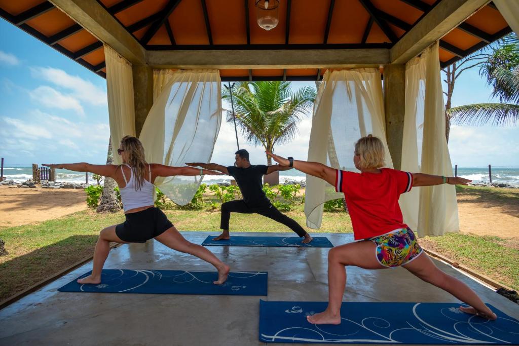 Jogas nodarbība brīvā dabā, kurā trīs cilvēki veic pozas zem klaja jumta ar skatu uz okeānu un palmas fona.