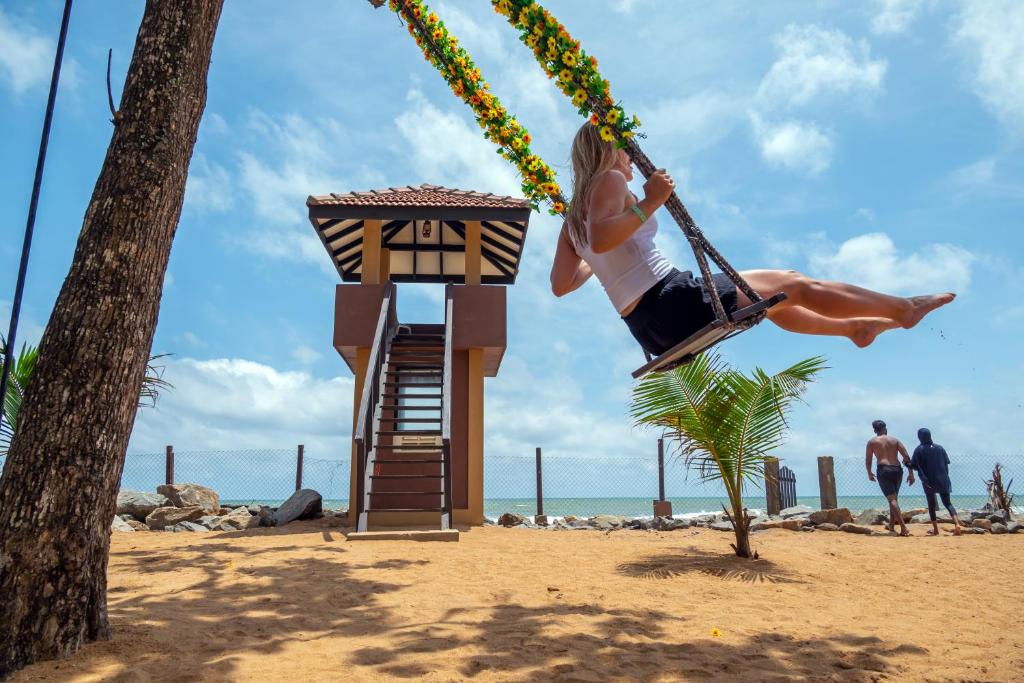 Šūpuļtīkls ar ziediem, kurā šūpojas sieviete pludmalē, blakus glābšanas tornim un jūrai, saulains laiks un smilšaina pludmale.