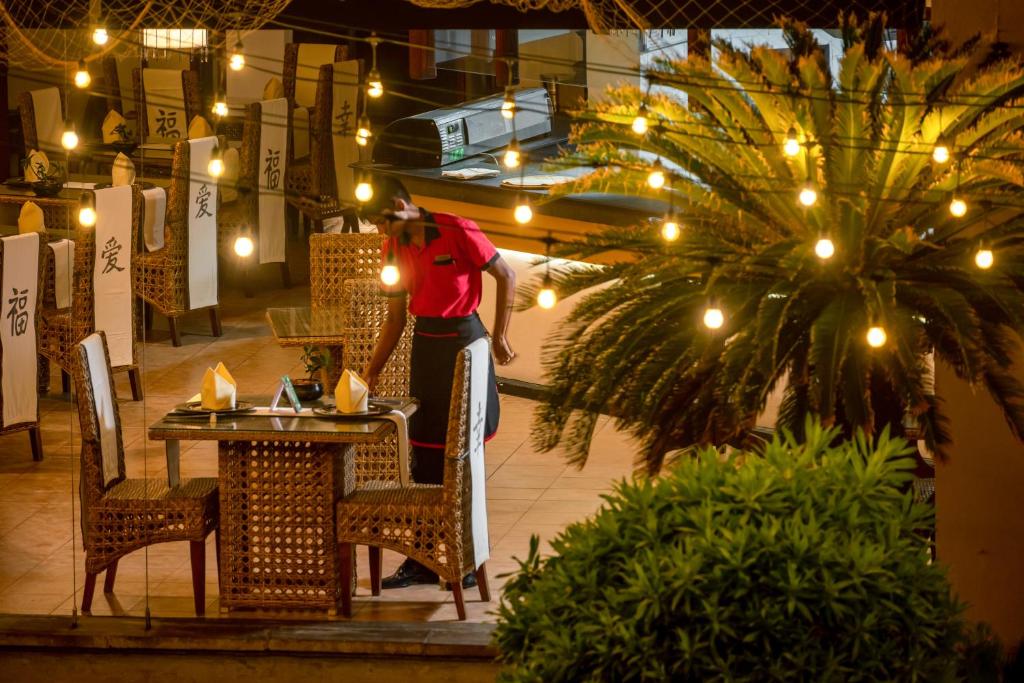 Restorāna interjers ar siltu apgaismojumu, kur apkalpotājs sagatavo galdu. Redzami ratiņi un tropiskie augi, radot patīkamu atmosfēru.