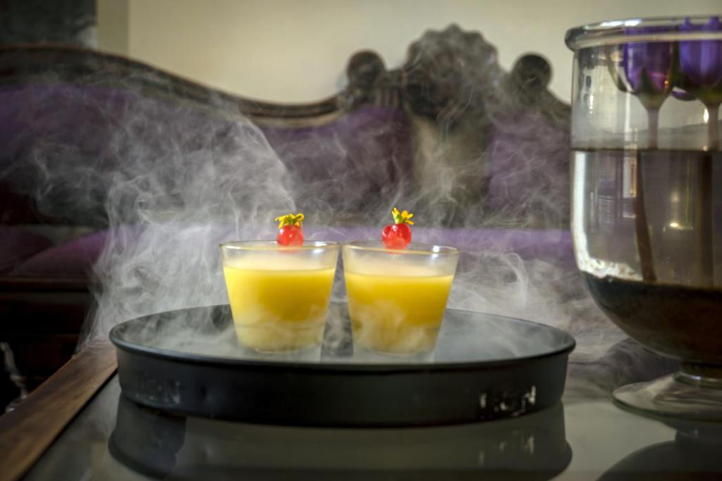 Divi dzērieni ar dūmiem un augļu dekoru, pasniegti uz melna šķīvja. Fons veido relaksējošu atmosfēru ar mīksto mēbelēm un ūdens elementiem.