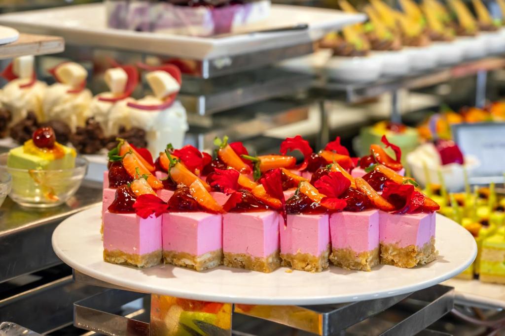 Saldējumā desertu plāksne ar rozā krāsas kūkām, rotātas ar augļiem un ziedlapiņām. Fons ar dažādiem citiem saldumiem.