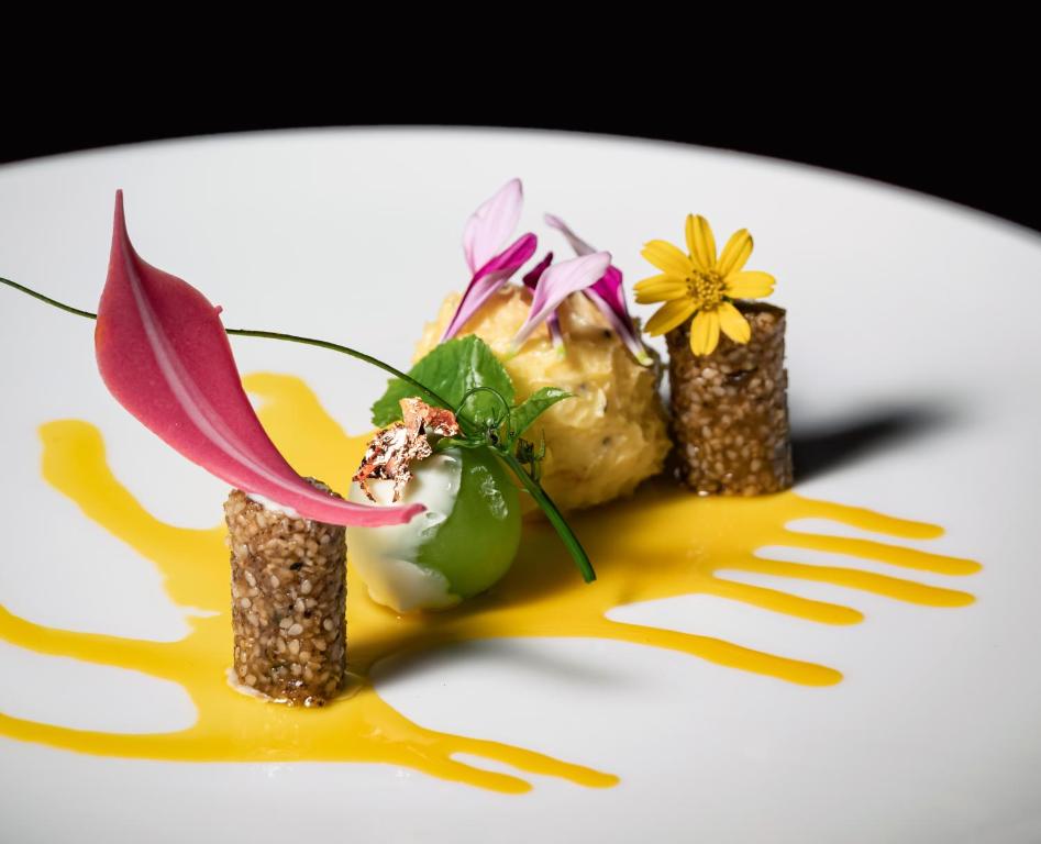 Eleganta deserts kompozīcija ar krāsainām sastāvdaļām, tostarp ziediem un augļu garnējumu, uz balta šķīvja. Izsmalcināts dizains ar krēmīgu pamatni un radošu dekorāciju.