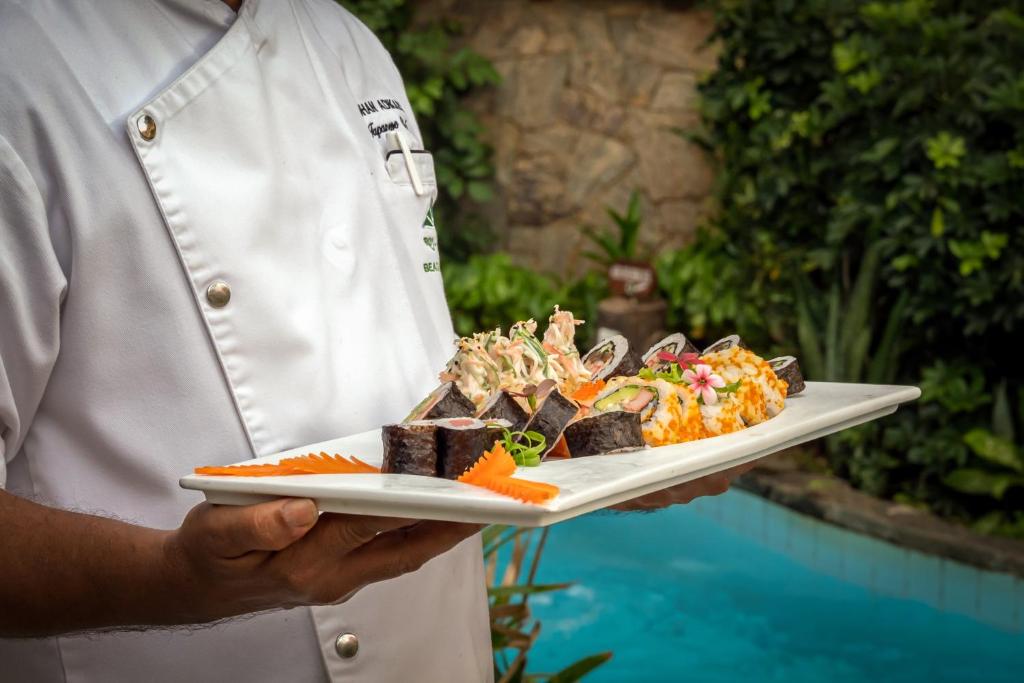 Šefpavārs pasniedz sushi uz baltas šķīvja, kas rotāts ar krāsainām garnelēm, avokado un dekoratīvu ziedu, fona veido zaļā dārzā esošs baseins.