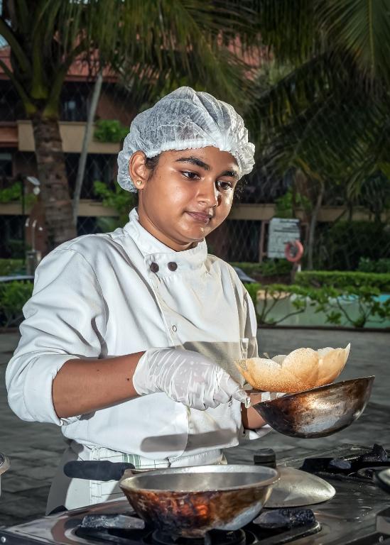Jaunais pavārs gatavo ēdienu ārā, valkājot baltu cepuri un aizsargcimdi. Apkārt ir zaļa vide un piepeldēšanas baseins.