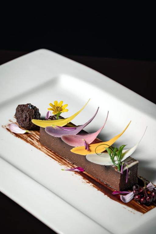 Dekorēts šokolādes deserts ar krāsainām lapām un ziediem, pasniegts uz eleganta balta šķīvja.