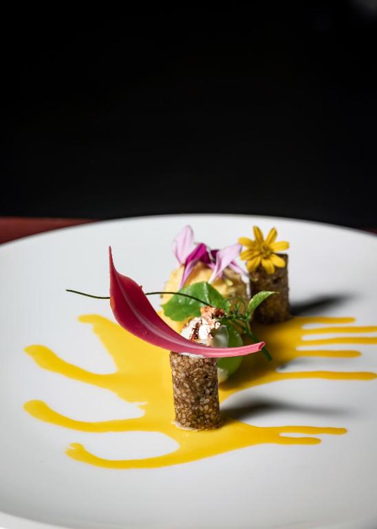 Elegantā plate ar mākslinieciski pasniegtu ēdienu, kas satur ziedus un krāsainu mērci. Radoša prezentācija un uzmanības pievēršana detaļām.