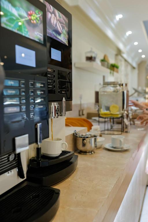 Kafijas automāti ar digitālajiem ekrāniem, kas sniedz iespēju pagatavot dažādus dzērienus, atrodas modernā kafejnīcā. Blakus ir glīti pasniegti kafijas krūzītes un ūdens dzesētājs ar citrona šķēlēm.