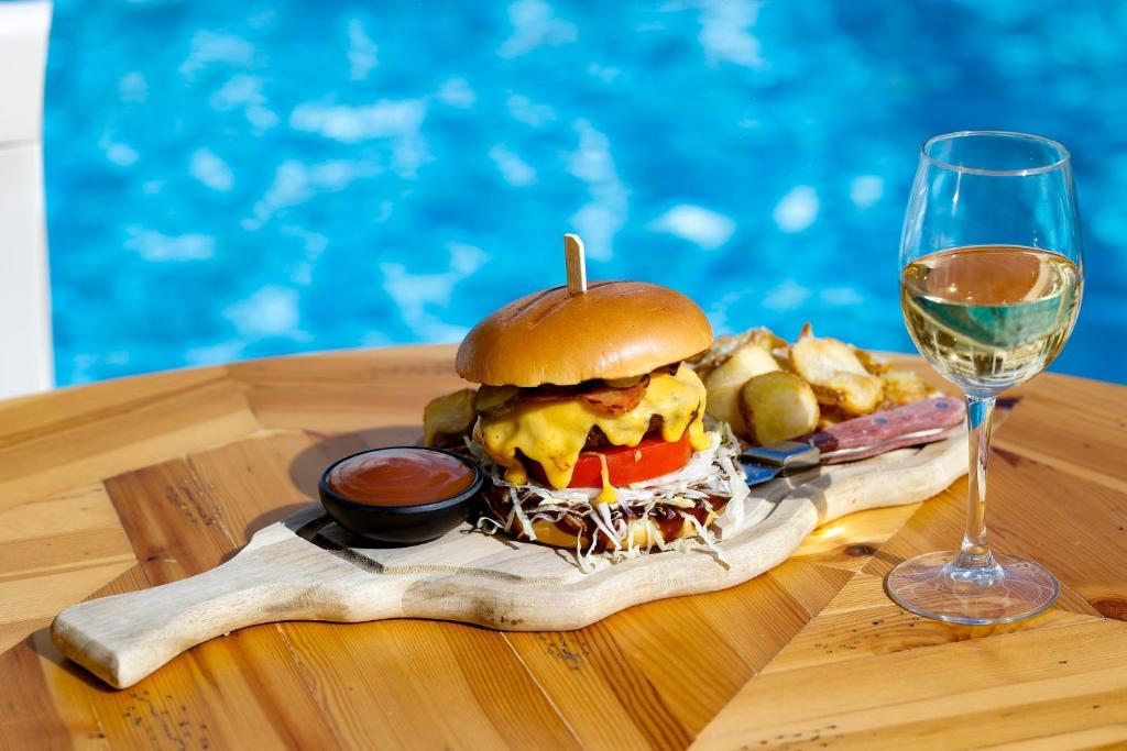 Garšīgs burgeris ar sieru un dārzeņiem, pasniegts uz koka dēlīša kopā ar mērci un ceptiem kartupeļiem, blakus stāv glāze balto vīna. Fons ar zilu ūdeni rada patīkamu atmosfēru.