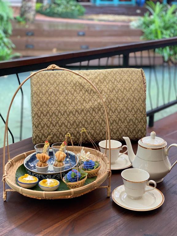 Elegants tējas servēšanas komplekts ar dažādiem desertiem un tējkannu, novietots uz koka galda. Fons veido zaļi dārzi, radot mierīgu atmosfēru.