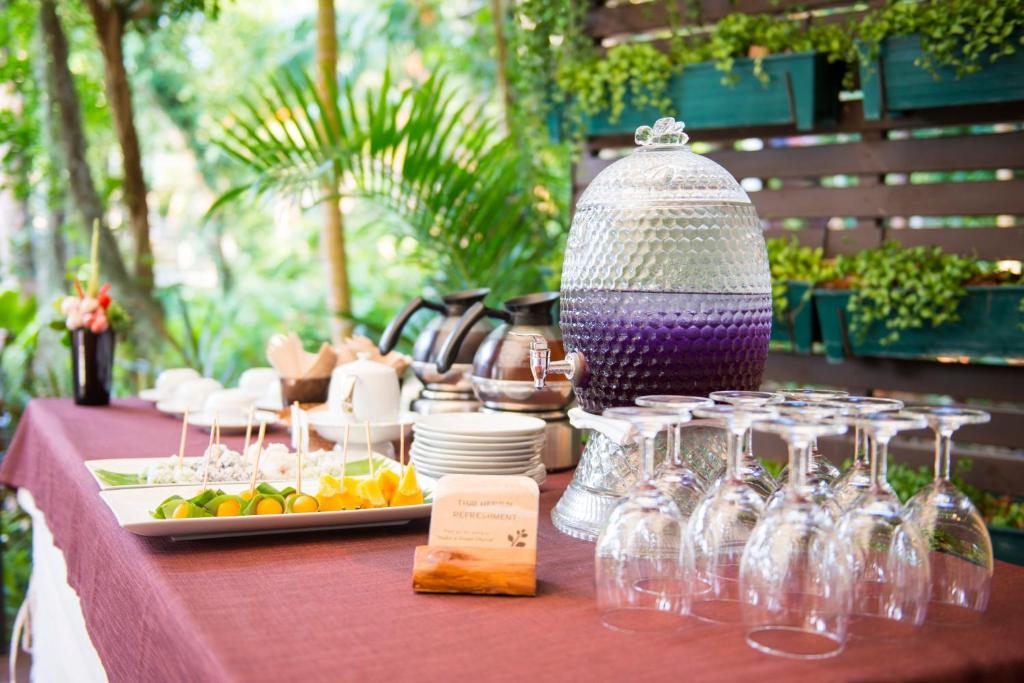 Apsveikumu galda klāšana ar dzērienu dozatoru un dažādiem uzkodu šķīvjiem, ieskaitot augļus un saldumus, zaļo augu fonā.