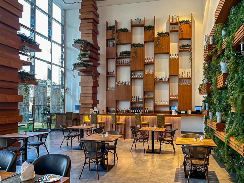 Moderns restorāns ar stilīgu interjeru, kas ietver koka elementus un zaļus augus. Bāra plaukts ir piepildīts ar dažādiem dzērieniem, radot patīkamu un relaksējošu atmosfēru.