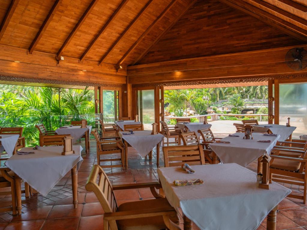 Koka interjera restorāns ar galdiem un krēslām, kas izkārtoti ar baltām galdautām, un skatu uz zaļojošiem augiem ārpus logiem.