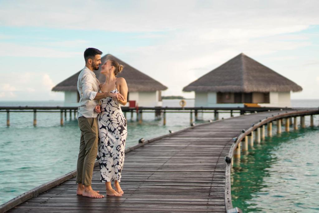 Romantiska pāris brīvdienās pludmalē, stāvot uz koka tilta pie ūdens, fona viesnīcu būves ar salmu jumtiem.