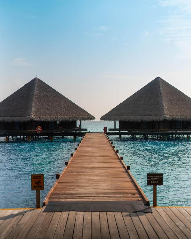 Koka laipa ved uz divām tropiskām viesnīcām ar salmu jumtiem, kas atrodas uz ūdens, ar skatu uz mierīgu jūru un zilo debesu fona.