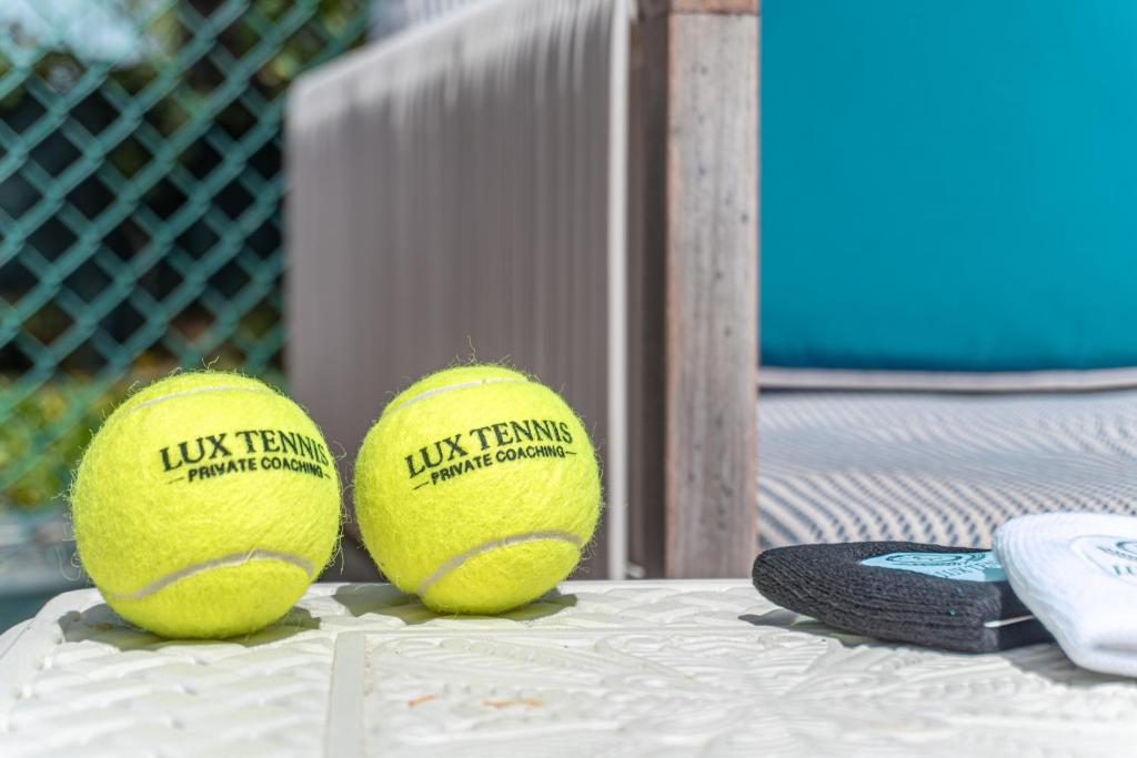 Divas dzeltenas tenisa bumbas ar uzrakstu "LUX TENNIS PRIVATE COACHING" un divi dvieļi uz galda, fons ar zaļu žogu un mīkstu mēbeļu.