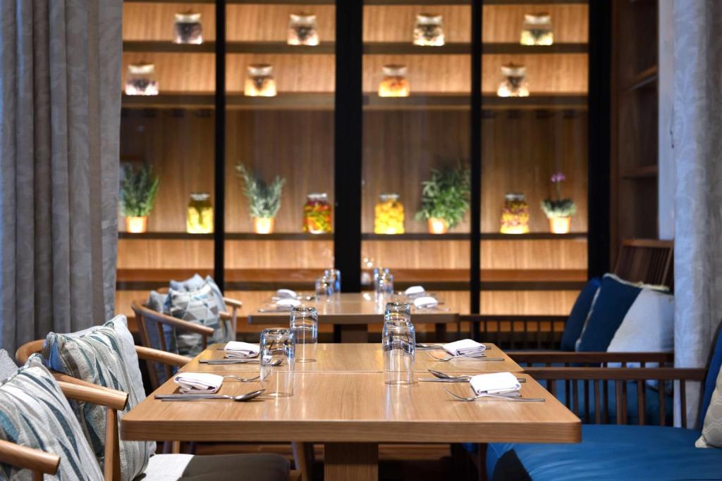 Mūsdienīgs restorāna interjers ar koka galdiem un ērtām mēbelēm, apgaismots ar krāsainām burkām uz plaukta. Uz galdiem redzami galda piederumi un glāzes.