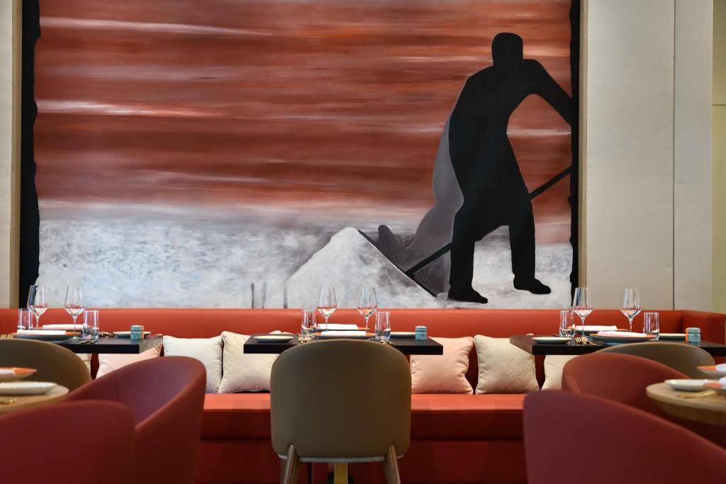 Mūsdienīgs restorāns ar sarkanu dīvānu un elegantiem galdiem, kam fonā ir mākslinieciska glezna ar melnu figūru un siltu krāsu paleti.