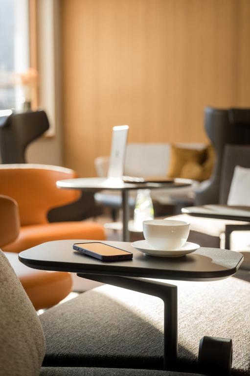 Mūsdienīgs kafejnīcas interjers ar ērtām atpūtas krēslām, uz galdiņa novietota tase ar kafiju un mobilais tālrunis. Dabiska apgaismojuma atmosfēra rada relaksējošu vidi.