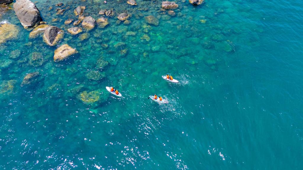 Trīs kajaki burā pa skaidro zilo ūdeni, apkārt redzami akmeņi un dabiski ūdens attēli. Ideāla ainava aktīvai atpūtai pie ūdens.