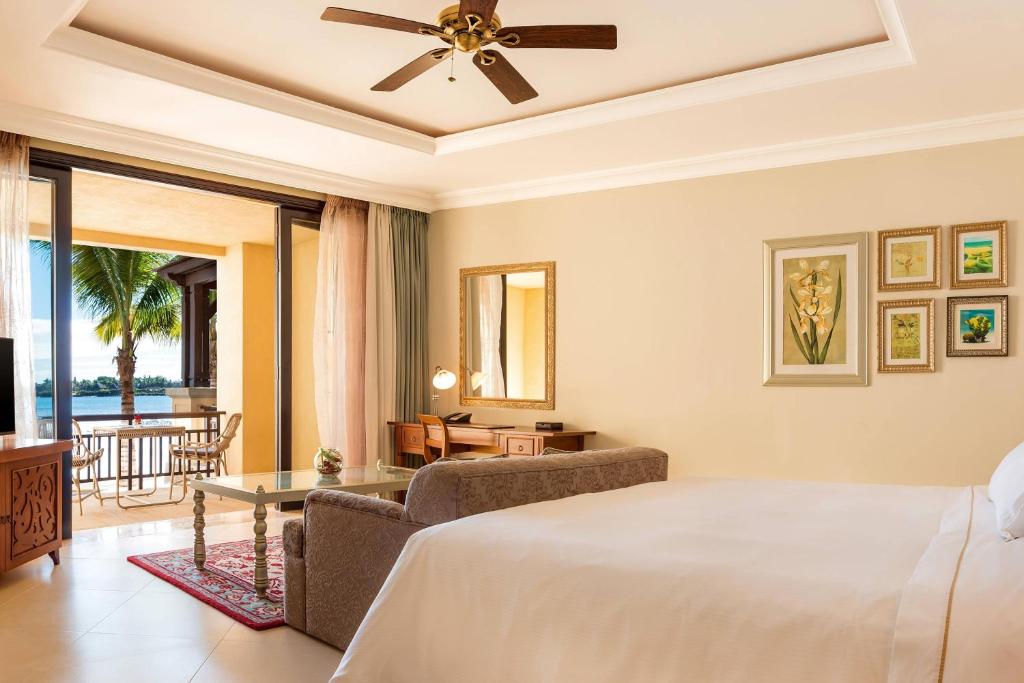 Elegants viesnīcas numurs ar skatu uz jūru, iekārtots ar mūsdienīgu mēbelēm un mākslas darbiem uz sienām. Lielas logu durvis ved uz terasi ar dārza mēbelēm un palmām.