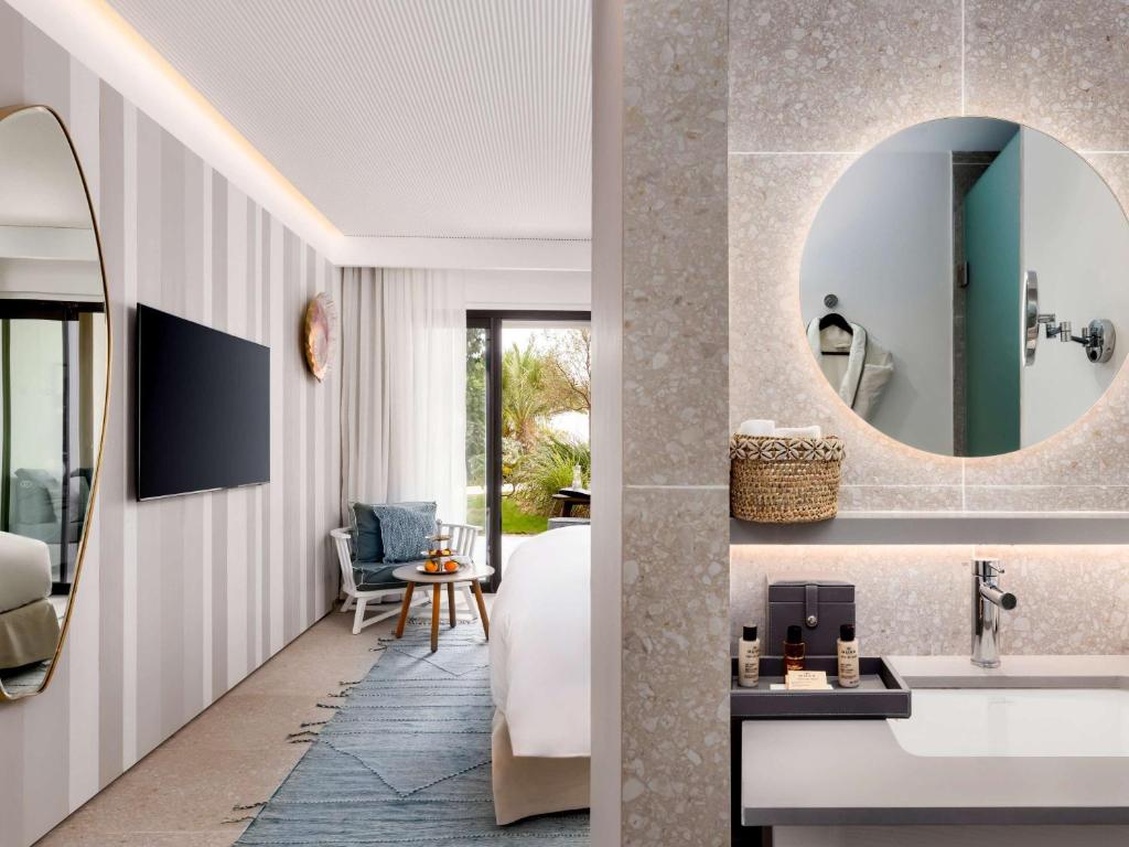 Mūsdienīgs viesnīcas numurs ar elegantu dizainu, kurā redzama dzīvojamā telpa ar televizoru un gultu, kā arī vannas istaba ar apaļu spoguli un moderno aprīkojumu. Izskatās gaisīgs un komfortabls.