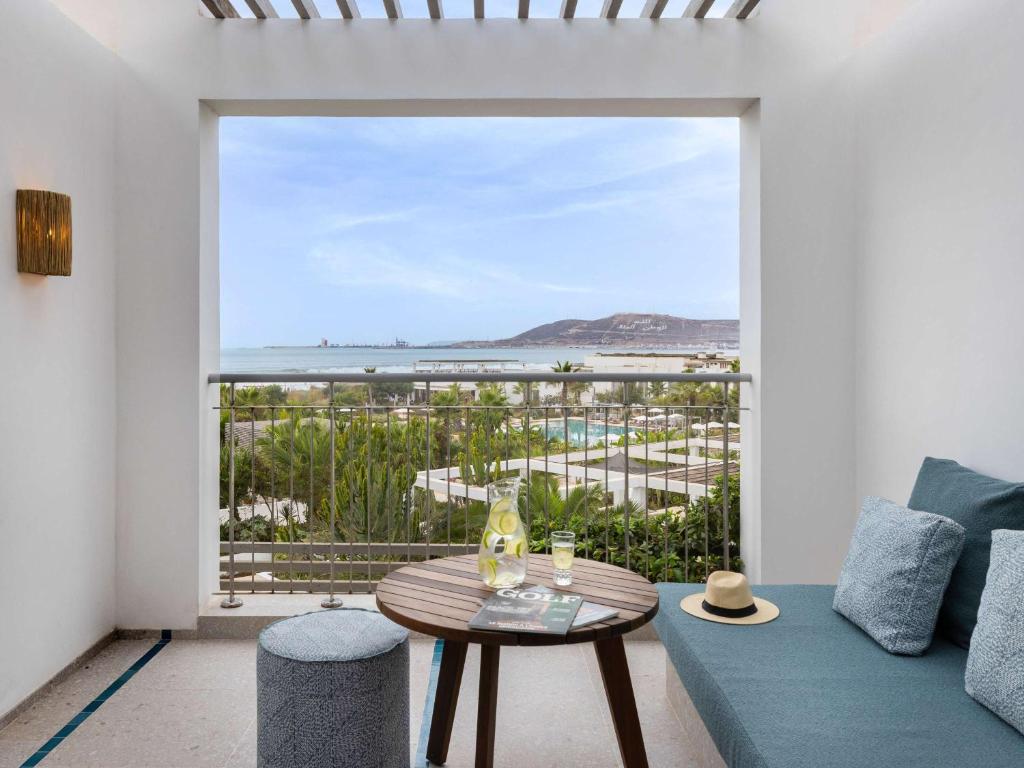 Balkons ar skatu uz jūru, kur atpūšas viesnīcas viesis, blakus ir galds ar dzērienu un žurnālu. Zaļie augi un baseins redzami lejā.