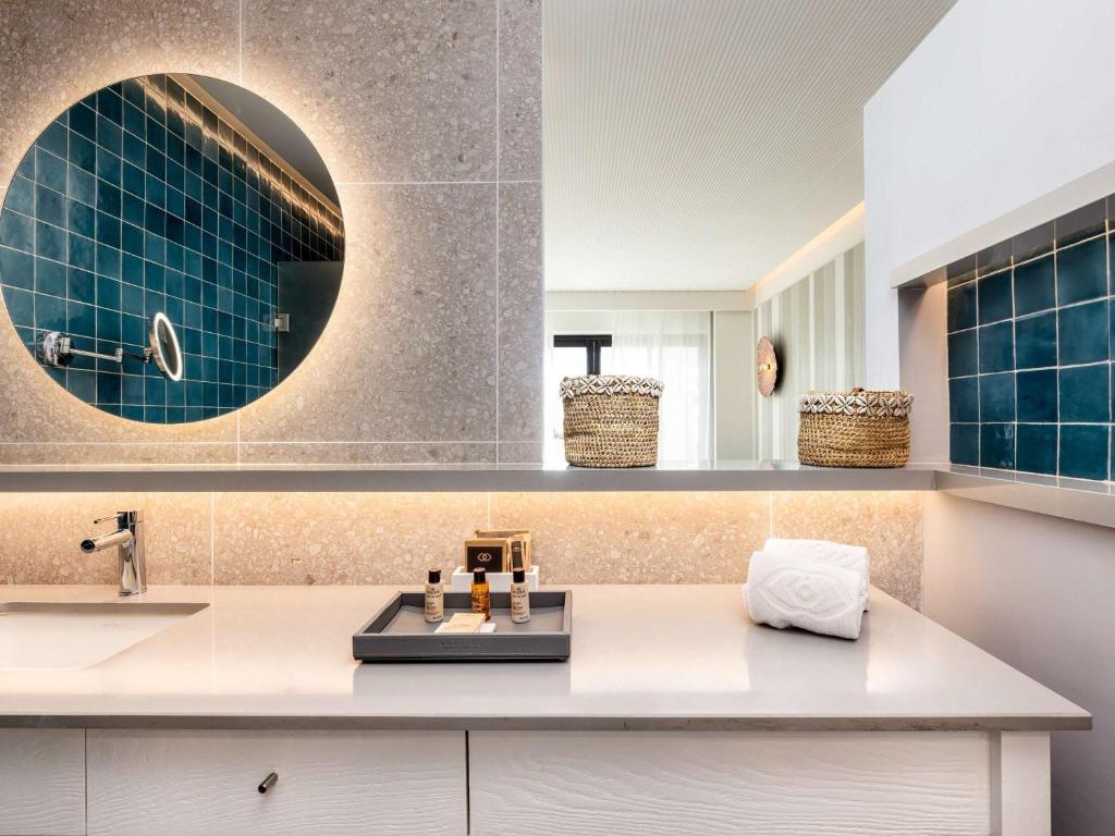 Mūsdienīgs vannas istabas interjers ar zilām flīzēm, apaļu spoguli ar apgaismojumu un elegantu izkārtojumu, kurā redzams trauks ar kosmētiku un dvieli.